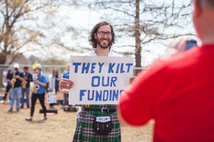 man holding a sign that reads "They Kilt our funding". He is also wearing a kilt