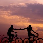 A backlit couple holding hands while biking