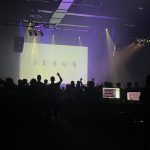 Picture of people worshipping in a dark room with the word "Jesus" displayed on the screen